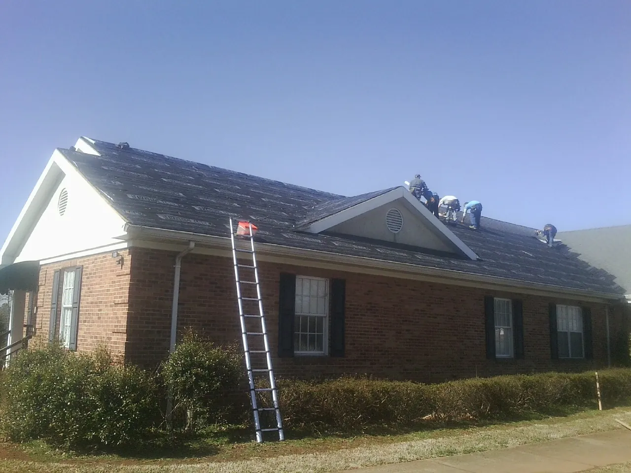 Crew tearing shingles off a brick ranch roof with a ladder set — Emergency Roof Repair work by our crew in Hay