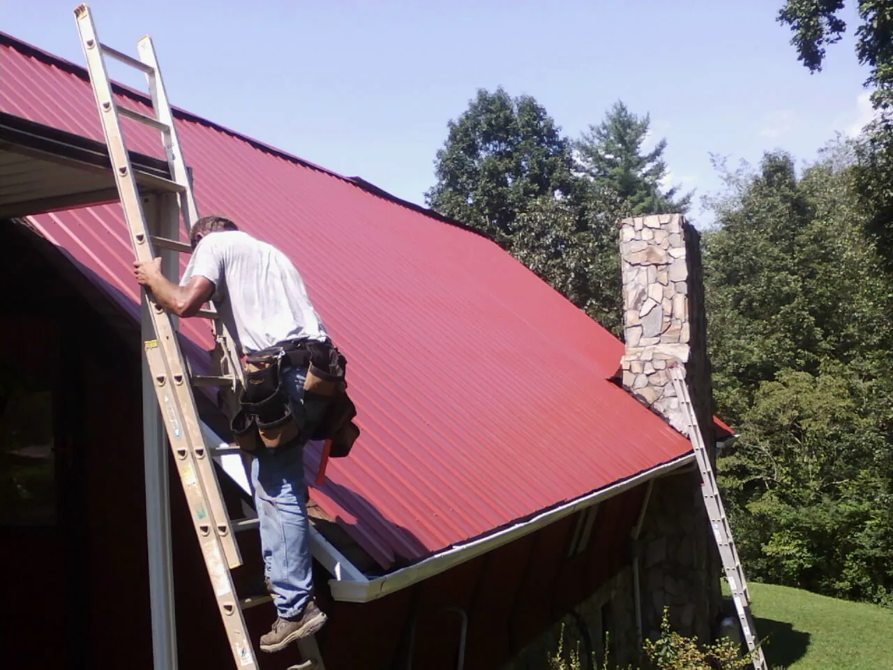 Roofer on a ladder beside a red metal roof and stone chimney — Soffit & Fascia Repair by Roofing Brothers in Hay